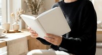 Woman in black sweater reading open book while sitting at wooden table with coffee cup in cozy home interior for relaxation and leisure activities.