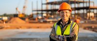 Confident construction worker in safety gear standing at building site during golden hour with industrial equipment and steel framework in background.