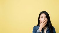 Surprised Asian woman with hand covering mouth wearing denim shirt against yellow background, expressing shock and amazement for advertising and marketing campaigns.