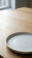 Empty white ceramic plate on light wood table near window with natural daylight, minimalist dining setup for restaurant, home decor, food styling.