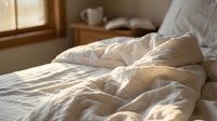 Unmade bed with white rumpled bedding and pillows in warm morning sunlight, suitable for lifestyle, home decor, and wellness content.