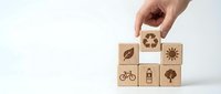 Hand placing wooden cube with recycling symbol among eco-friendly icons including bicycle, tree, sun, and water bottle on white background.
