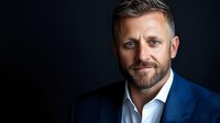 Confident mature Caucasian businessman with beard wearing blue suit and white shirt against dark background, professional corporate headshot portrait with dramatic lighting.