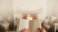 Young woman with light skin tone reading open book near fireplace with warm glowing flames and cozy home interior setting for relaxation and literature themes.