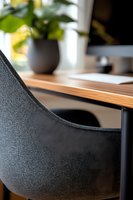 Modern workspace detail showing black textured office chair and wooden desk surface with potted plant and computer monitor in soft natural lighting.
