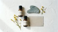 Natural skincare products with essential oil bottles, gua sha stone and soap bar arranged with dried flowers on white textured background for wellness branding.