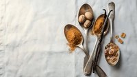 Vintage spoons filled with cocoa powder and whole nutmeg on white marble surface for baking and cooking recipes.
