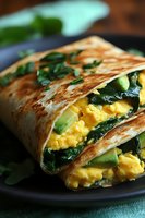 Grilled breakfast wrap filled with scrambled eggs, fresh avocado and spinach leaves on dark plate, close up view showing layers of morning meal ingredients.