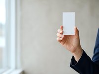 Caucasian male hand holding blank white card against neutral wall background with natural window light, suitable for business card mockups, corporate branding, and marketing presentations.