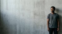Young man with dark hair and medium skin tone wearing casual gray t-shirt standing against weathered concrete wall, suitable for lifestyle, fashion, and portrait projects.