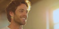 Smiling young Caucasian man with tousled brown hair and stubble in warm sunlight, creating positive atmosphere for lifestyle and wellness marketing.