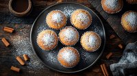 Freshly baked vanilla muffins dusted with powdered sugar on dark plate, scattered cinnamon sticks on rustic wooden table, overhead view with moody lighting.