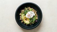 Overhead view of a gourmet grain bowl with quinoa, roasted vegetables, microgreens, and a poached egg with runny yolk on black plate. Food styling and restaurant presentation.