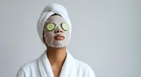 Young woman enjoying spa treatment with cucumber slices on eyes and facial mask, wearing white bathrobe and towel turban for skincare routine.