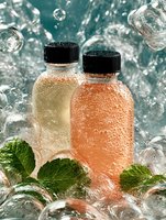 Refreshing bottles of sparkling beverages with mint leaves surrounded by water splashes and ice cubes on turquoise background.