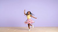 Young girl with long dark hair and medium skin tone, wearing yellow top and pink patterned skirt, dancing joyfully with arms raised on wooden floor against purple background.