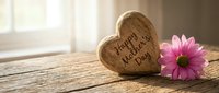 Wooden heart with Happy Mother's Day text and pink daisy flower on rustic table in warm sunlight for greeting card design.