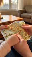 Child holding colorful road map activity book with illustrated streets and vehicles indoors near wooden table and beige armchair by sunlit window for educational play and entertainment.