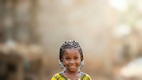 Young African girl with braided hairstyle wearing colorful traditional clothing, smiling at camera with blurred background, suitable for diversity, culture, and childhood themes.
