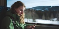Young woman in green jacket using smartphone while traveling by train, with snowy winter forest landscape visible through window.