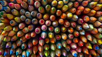 Vibrant colored pencils arranged in tight rows creating abstract pattern, shot from above showing sharp tips in rainbow hues against dark background.