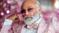 Senior Caucasian man with white beard wearing pink bow tie and patterned shirt poses confidently against pink bokeh background, expressing joy and style.