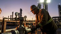 Industrial worker in safety gear operating valve equipment at oil refinery during sunset with dramatic lighting and industrial infrastructure background.