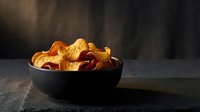 Golden crispy potato chips in black bowl on dark wooden table with dramatic lighting and smoky atmosphere for snack food advertising.