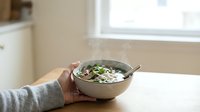 Asian noodle soup with vegetables and herbs served in ceramic bowl, held by person in casual setting for food photography and culinary content.