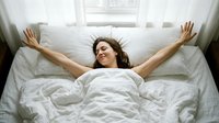Woman with dark hair and light skin tone stretching arms overhead while lying in bed with white linens, expressing morning awakening, relaxation, and wellness for lifestyle and health content.