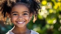 Young African American girl with natural curly hair and bright genuine smile outdoors in sunlit garden setting, radiating joy and innocence against blurred green background.