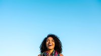 Young woman with curly dark hair and warm smile looking upward against clear blue sky, expressing joy and optimism for lifestyle and wellness content.