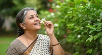 Mature woman of South Asian descent smelling white flowers in lush garden, wearing patterned top, enjoying nature and wellness outdoors.