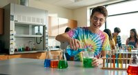 Young Caucasian male student conducting colorful chemistry experiment in school laboratory with test tubes and beakers for educational science learning.