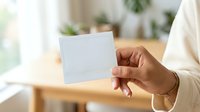 Young adult with medium skin tone holding blank white card in bright indoor workspace with plants and natural light, suitable for business, greeting, or promotional messaging.
