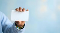 Young man with medium skin tone wearing light blue shirt holding blank white card covering face against soft blue background for messaging and communication concepts.