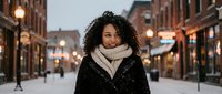 Young African American woman with curly hair wearing winter coat and scarf smiling on snowy city street at dusk with warm lights and historic buildings.