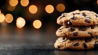Fresh baked chocolate chip cookies stacked on dark surface with warm glowing bokeh lights in background, creating cozy atmosphere for food photography.