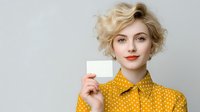 Young woman with short blonde curly hair wearing yellow polka dot shirt holding blank business card against light gray background. Perfect for business identity mockup.