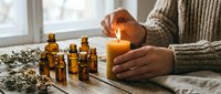 Cozy aromatherapy scene with essential oil bottles and burning candle on wooden table. Woman in knit sweater creating relaxing spa atmosphere at home.