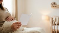 Young woman of mixed ethnicity wearing a beige knit sweater holding a blank white card in her hands while sitting indoors with warm lighting.