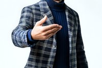 Young Asian man in plaid blazer and navy turtleneck sweater making welcoming hand gesture against white background, business casual style for modern professional.