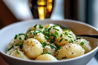 Creamy baby potatoes garnished with fresh chives and black pepper in white ceramic bowl, soft bokeh background creates warm restaurant ambiance.