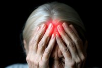 Senior woman covering face with hands, red glowing spot indicating headache, migraine or mental stress against dark background. Medical concept showing pain and suffering.
