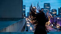 Young woman with flowing hair silhouetted against illuminated city skyline at night from rooftop terrace creating dramatic urban lifestyle moment.