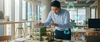 Male architect or designer in blue shirt reviewing sustainable building model in modern office with natural light and wooden furniture.