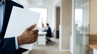 Close-up of male professional holding blank white document in modern office corridor with blurred colleagues in background, suitable for business communication and corporate concepts.