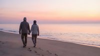 Romantic couple walking hand in hand on beach at sunset with soft pink and orange sky reflecting on calm ocean waves creating peaceful evening atmosphere.