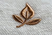 Golden leaf embroidery design on white fabric texture, delicate needlework detail showing metallic thread stitching in natural motif pattern.