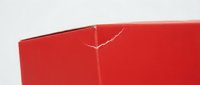 Red cardboard box corner showing white crack damage on folded edge against light background for packaging and shipping concepts.